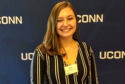 Class of 2021 senior Jenna Racca smiles in front of a UConn banner.