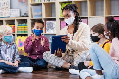 Kids and teacher wearing mask sitting on floor in classroom.