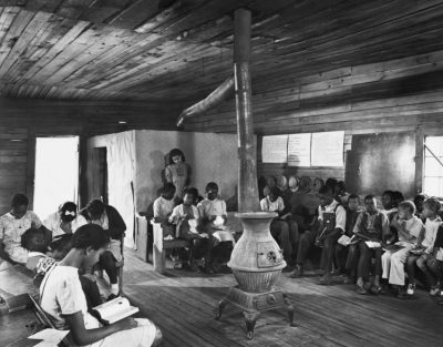 African American children sit in one-room school house around heater in a historic photo.