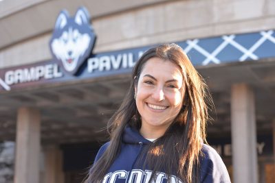 Smiling female with UConn shirt standing in front of the Gampel Pavillon. 