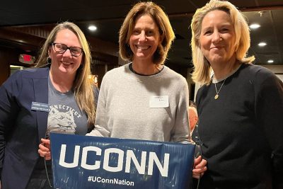 Three smiling woman hold a UConn banner at a reception.