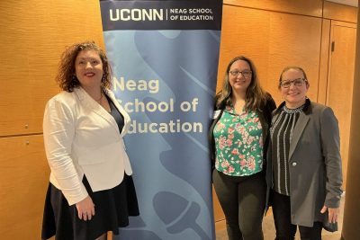 Three smiling female professionals stand next to Neag School banner.