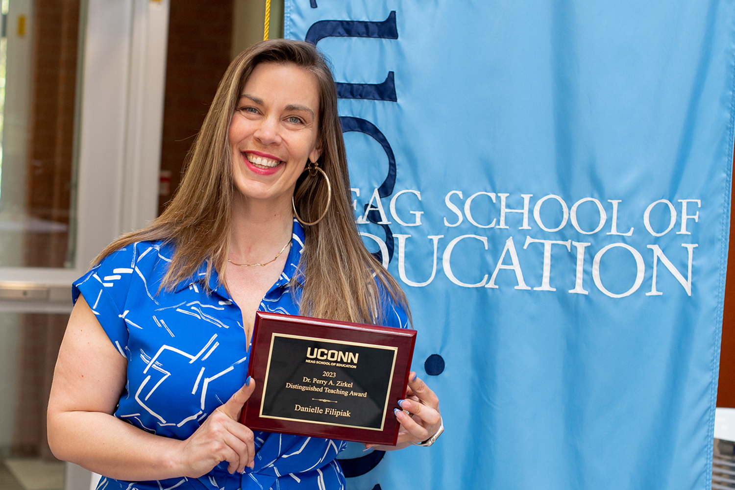 Danielle Filipiak holds her Zirkel award plaque while standing in front of the light blue Neag School of Education academic banner.