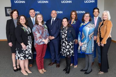Group of males and females wearing professional attire while standing in front of blue UConn banner.
