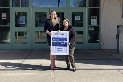 Two female educators stand in front of a school while holding a sign "Rogers Innovation Fund."