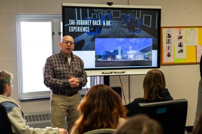 A white man stands in front of a projector screen and speaks to high school students.