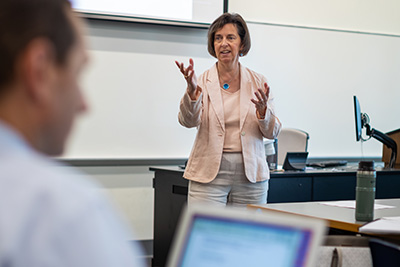 A white woman stands at the front of a classroom with a male student on a laptop in the foreground.