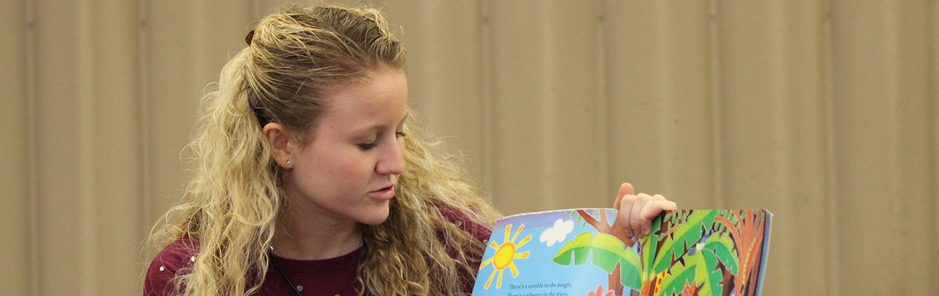 A white female student teacher reads a book out loud to students off screen.