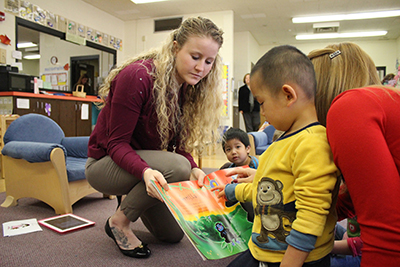 A white female student teacher helps an elementary student identify word sounds and story components in a picture book.