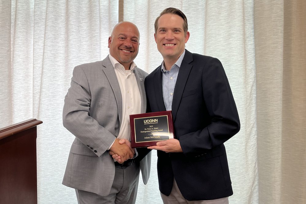 Two adult men pose and smile at the camera. One man is holding a small award plaque.