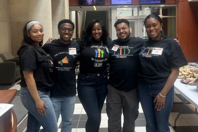 Five Black adults stand in a row and smile at the camera.