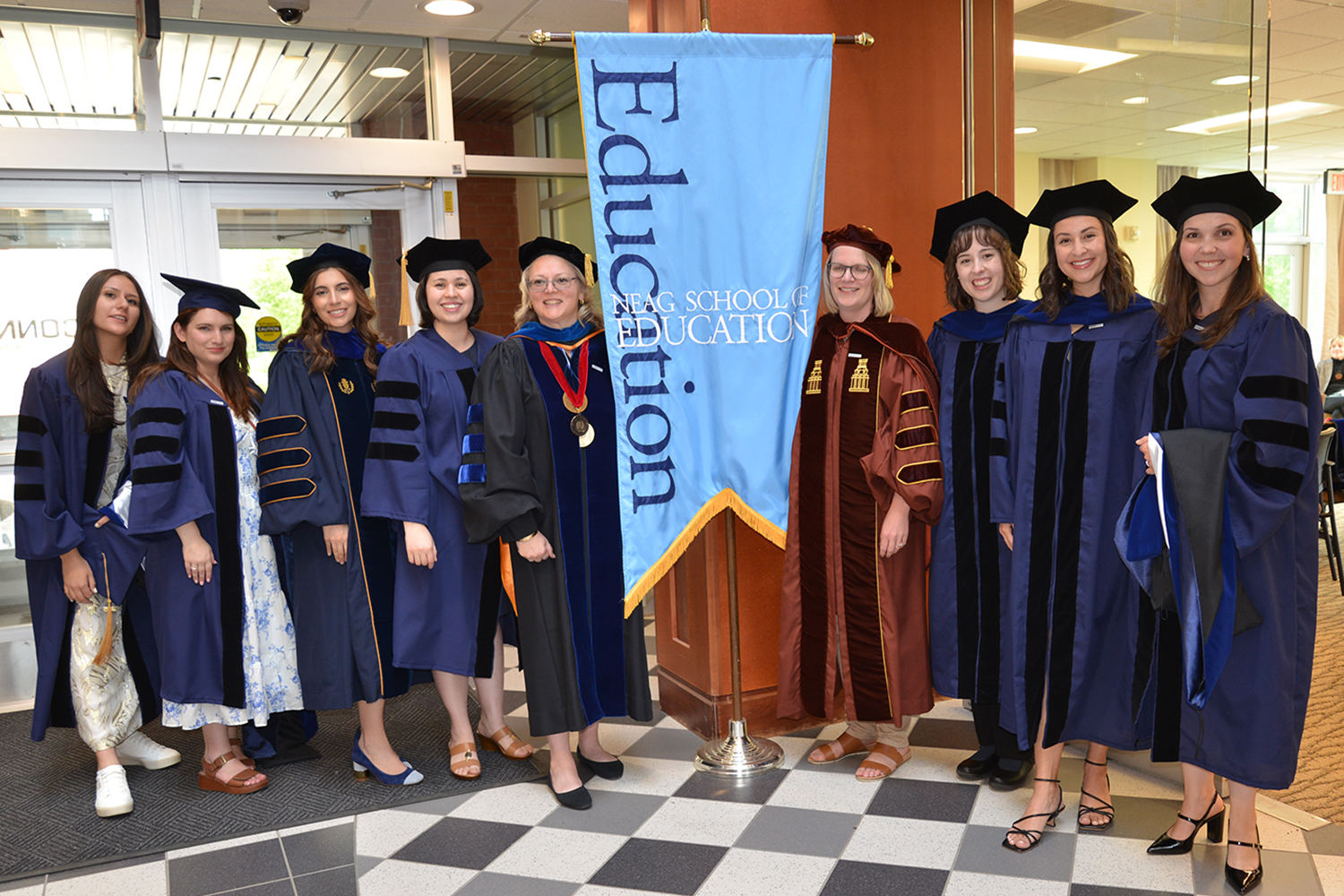 Nine women in doctoral robes stand next to a light blue UConn Neag School of Education banner.