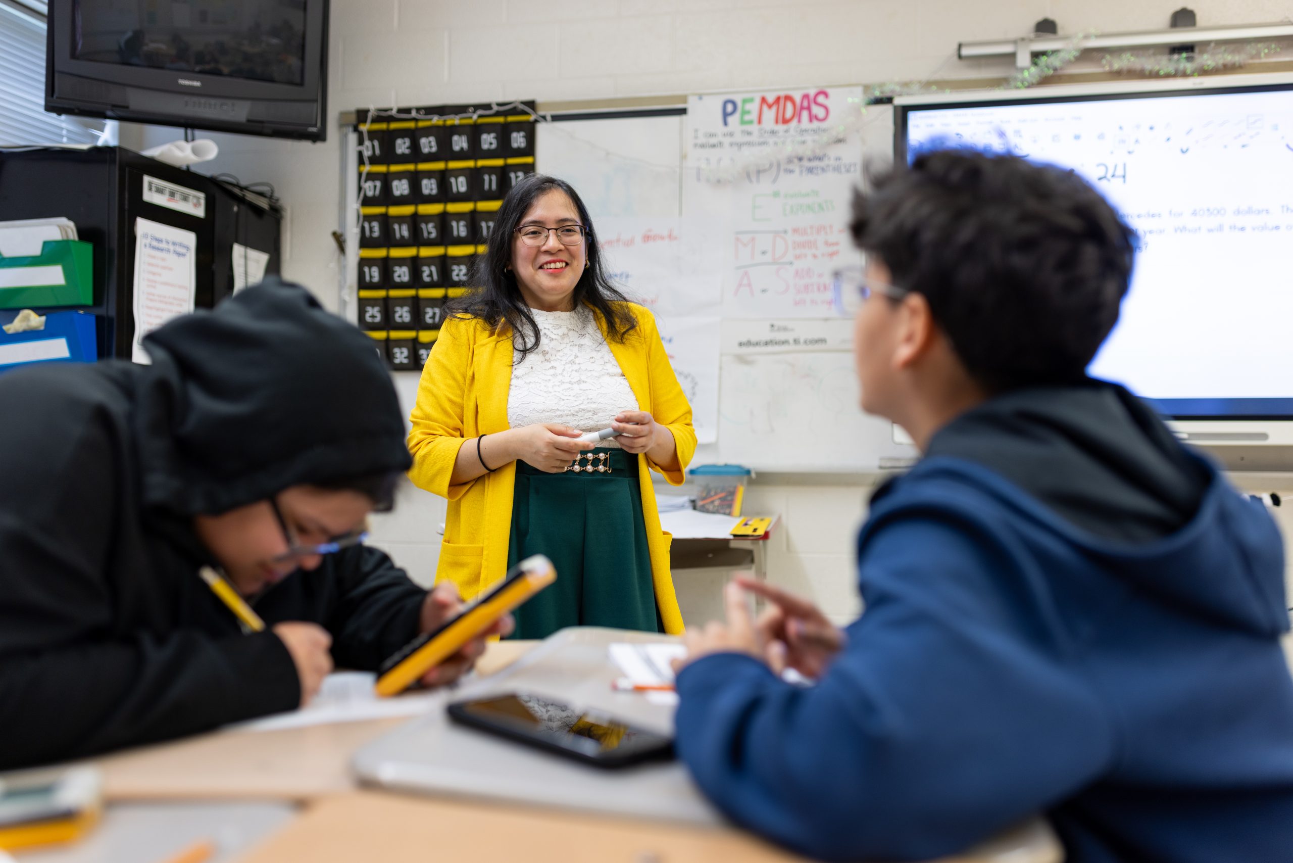 A female teacher stands in a classroom, wearing a yellow blazer and glasses, and smiles at a male student, who is sitting with their back to the camera.