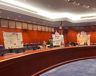 Three high school students sit in a legislative chamber.