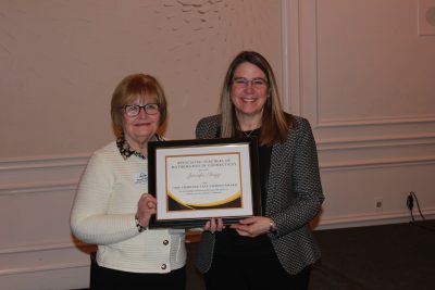 Two women hold a framed award certificate and smile at the camera.