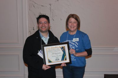A man and a woman hold a framed award certificate and smile at the camera.