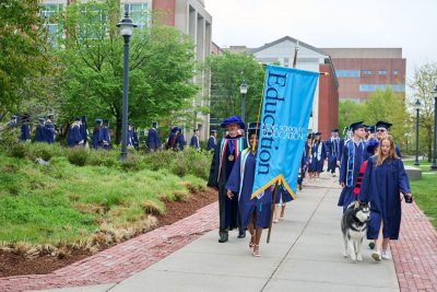 Graduates walk outside in two lines led by a light blue banner.