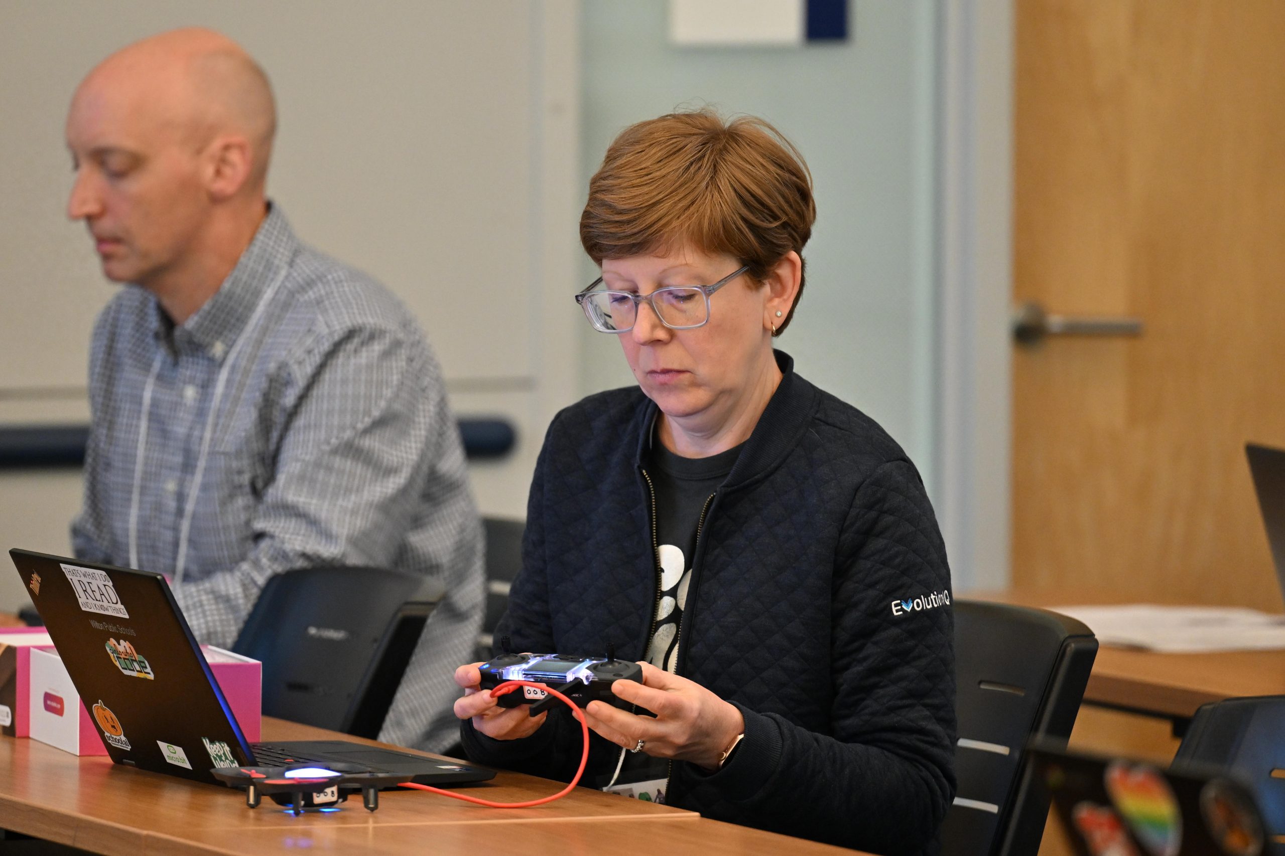 A female educator sits in a classroom and holds a piece of technology. A male educator sits next to her. [Links to event registration website].