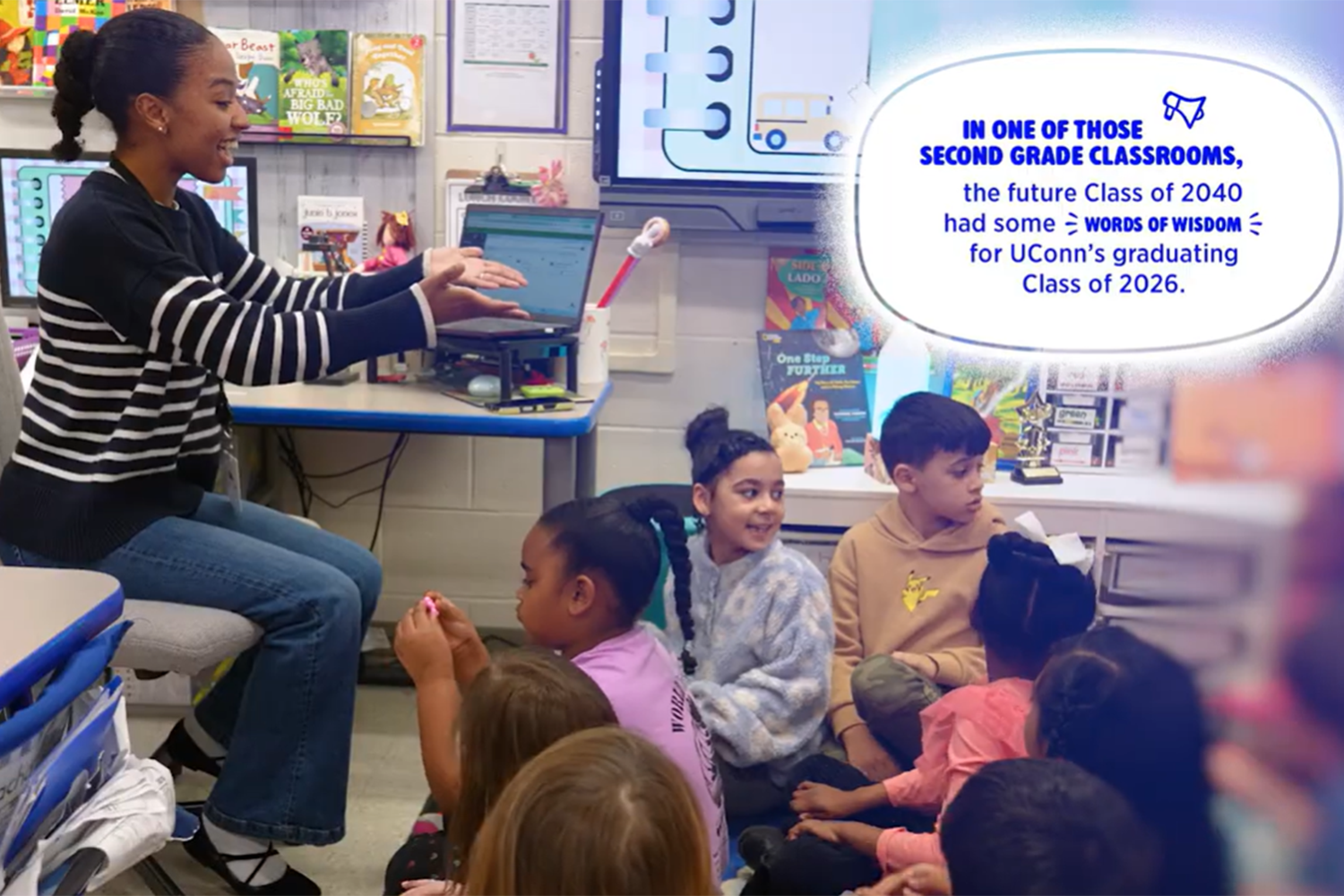 A female student teacher sits in a chair in a classroom, while elementary school students sit on the floor in front of her. [Links to YouTube video]
